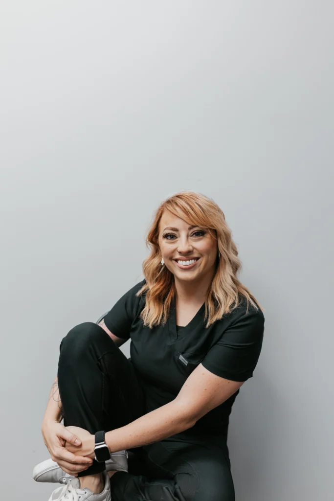 Smiling female dental assistant seated in black scrubs on a neutral background